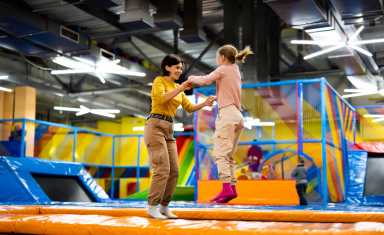 Pretty girl and her daughter kid jumping on colorful trampoline at playground park and smiling. Beautiful family during active entertainments