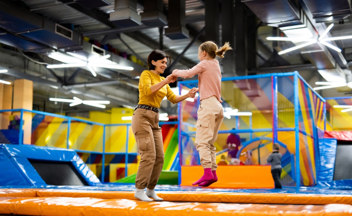 Pretty girl and her daughter kid jumping on colorful trampoline at playground park and smiling. Beautiful family during active entertainments