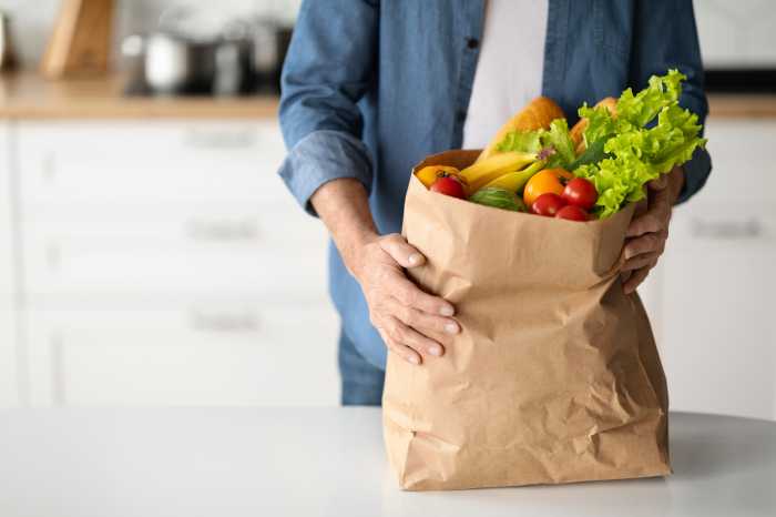 Healthy Nutrition Concept. Mature Man Holding Paper Bag With Groceries In Kitchen Interior, Cropped Shot Of Unrecognizable Senior Male Placing Pack With Fresh Organic Vegetables And Fruits On Table