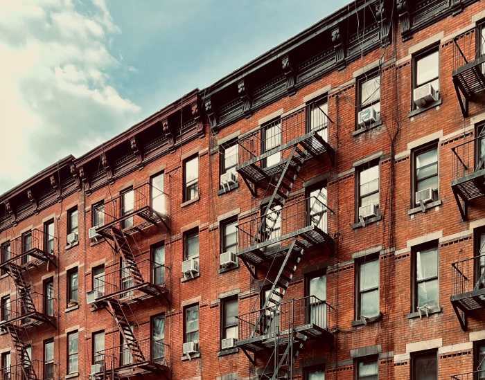 This image showcases a timeless urban scene of a brick apartment building adorned with iconic fire escapes. The warm tones of the brick contrast beautifully with the soft blue sky, capturing the essence of city life in a historical neighborhood. The architectural details, including the ornate cornices and window air conditioning units, evoke a sense of nostalgia and urban charm. Perfect for projects related to city living, historical architecture, or urban photography