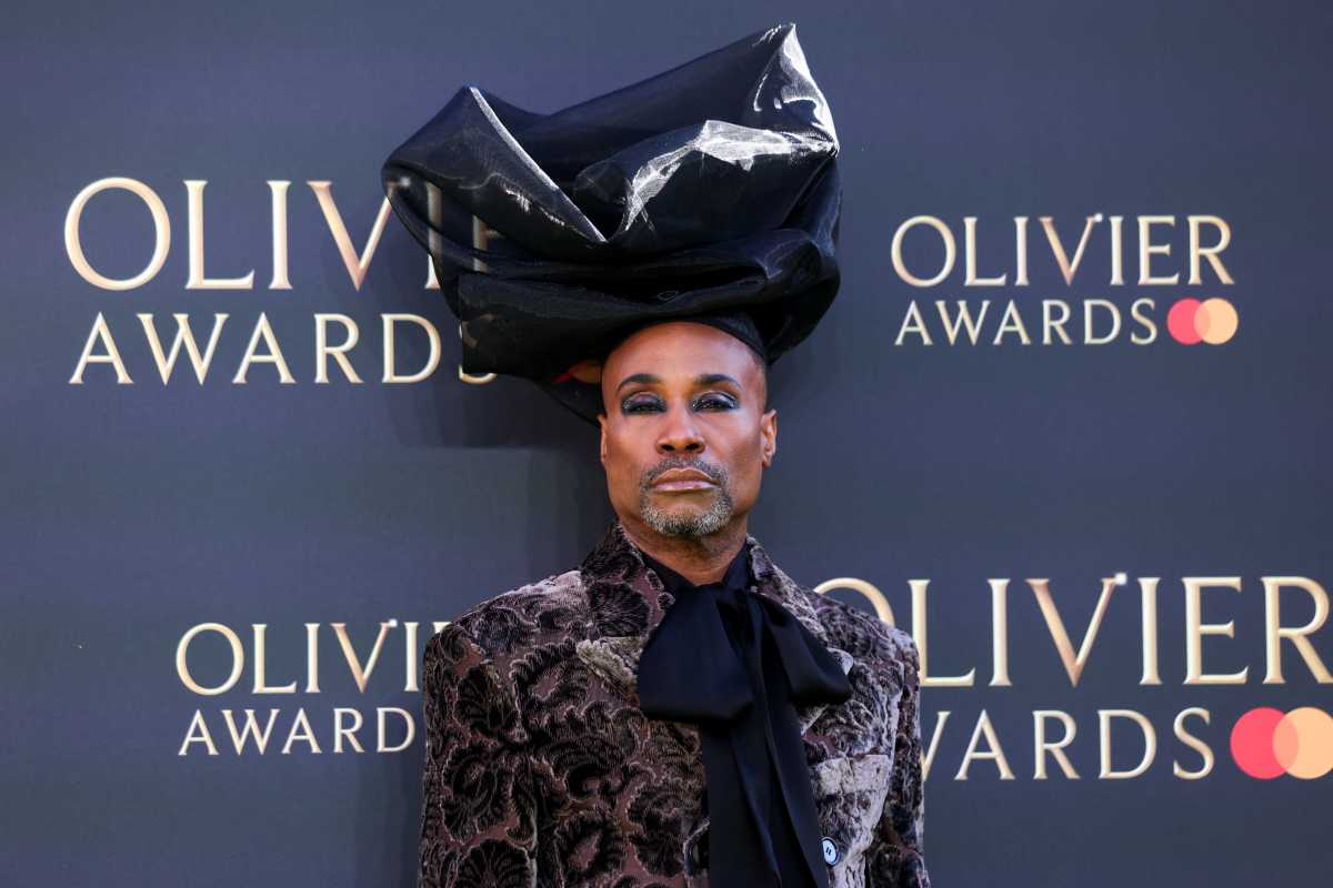 man wearing makeup, black headdress and velvet suit in front of Olivier Awards step-and-repeat