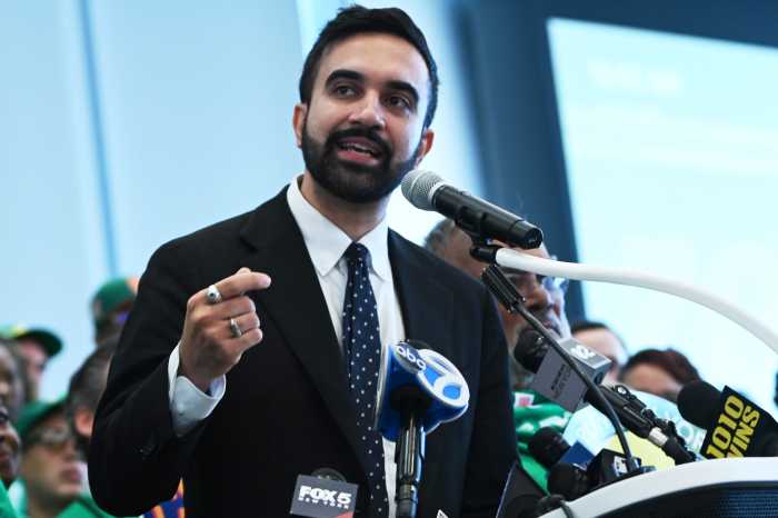 man with beard and suit Mamdani speaking at podium with microphones in front of him