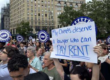 An advocate holds up a sign reading "My clients deserve a lawyer who can pay rent"