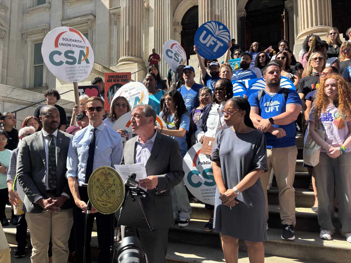 Man in suit stands in front of crowd holding signs and speaks to press.