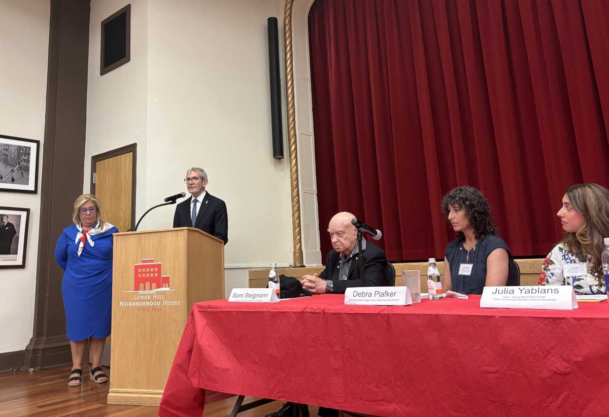 Man in a suit speaks at a podium next to a woman in a blue dress in front of a red curtain and next to a table of panelists.