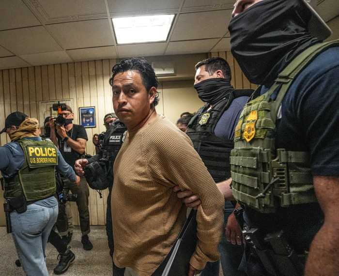 Man in beige shirt looks concerned while being arrested by masked ICE agent