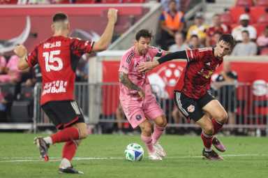 Lionel Messi: Man in pink uniform dribbles soccer ball between two men in red uniforms