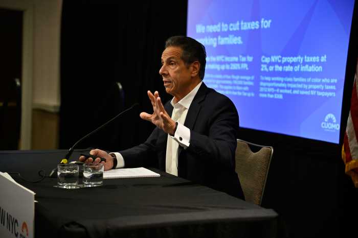 Man in a suit presents in front of a big screen, sitting at a table with a microphone