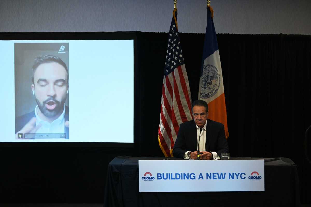 man in suit at desk next to video fo another man andrew cuomo and zohran mamdani