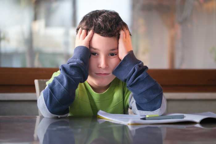 Little boy at home sitting at the table doing his homework by himself. Holding head by hand and sadly looking at the camera.