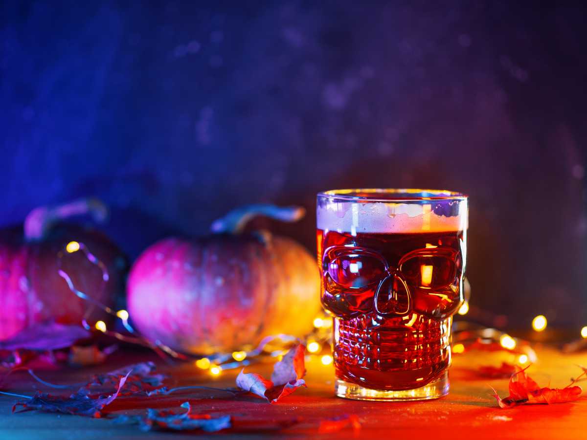 Red ale beer in a skull-shaped glass on a table with pumpkins and autumn leaves. Celebrate Halloween, copy space