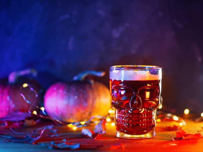 Red ale beer in a skull-shaped glass on a table with pumpkins and autumn leaves. Celebrate Halloween, copy space
