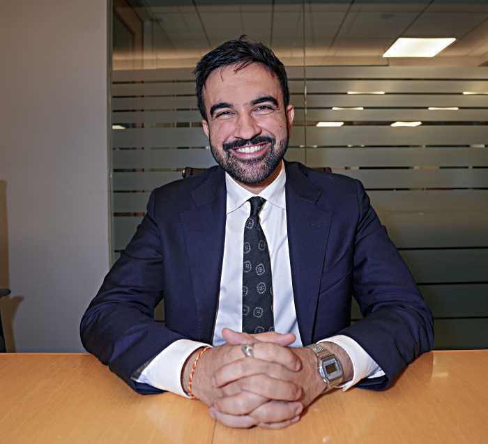 Zohran Mamdani smiling with hands folded in front of him