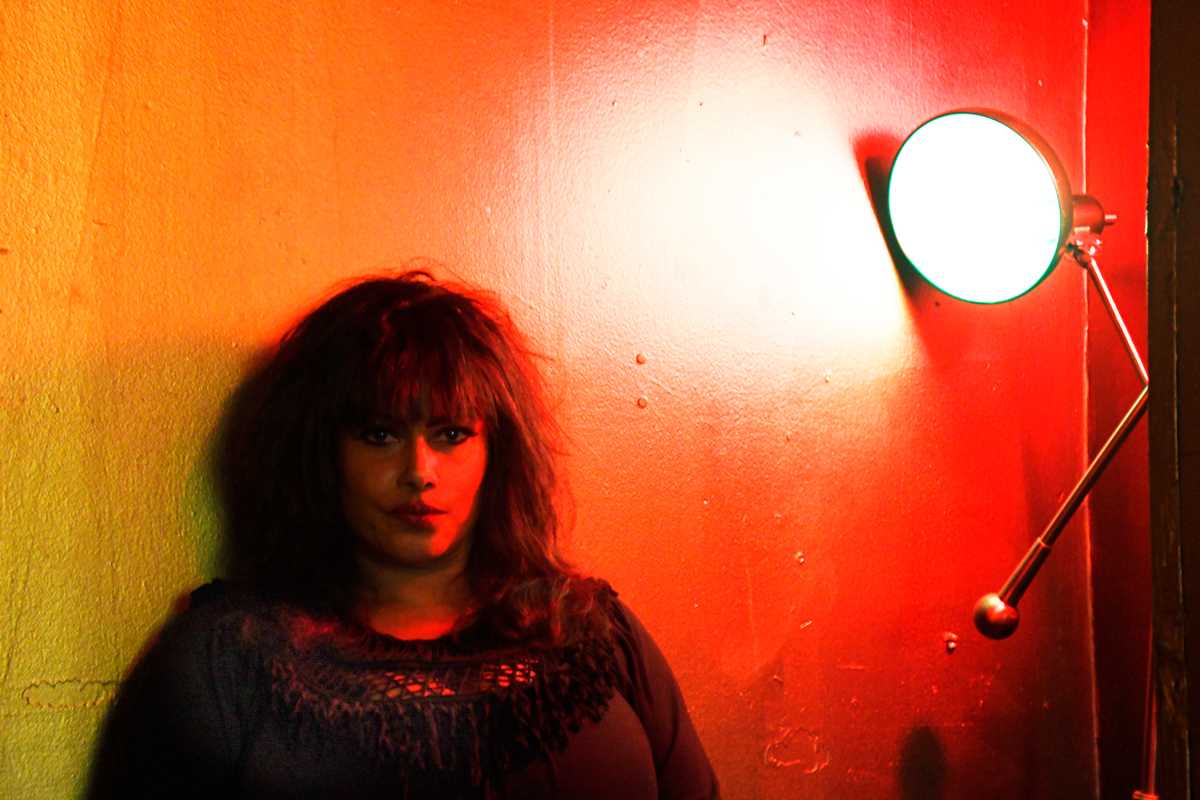 Sylvia Black in front of red wall with light at East Village club.