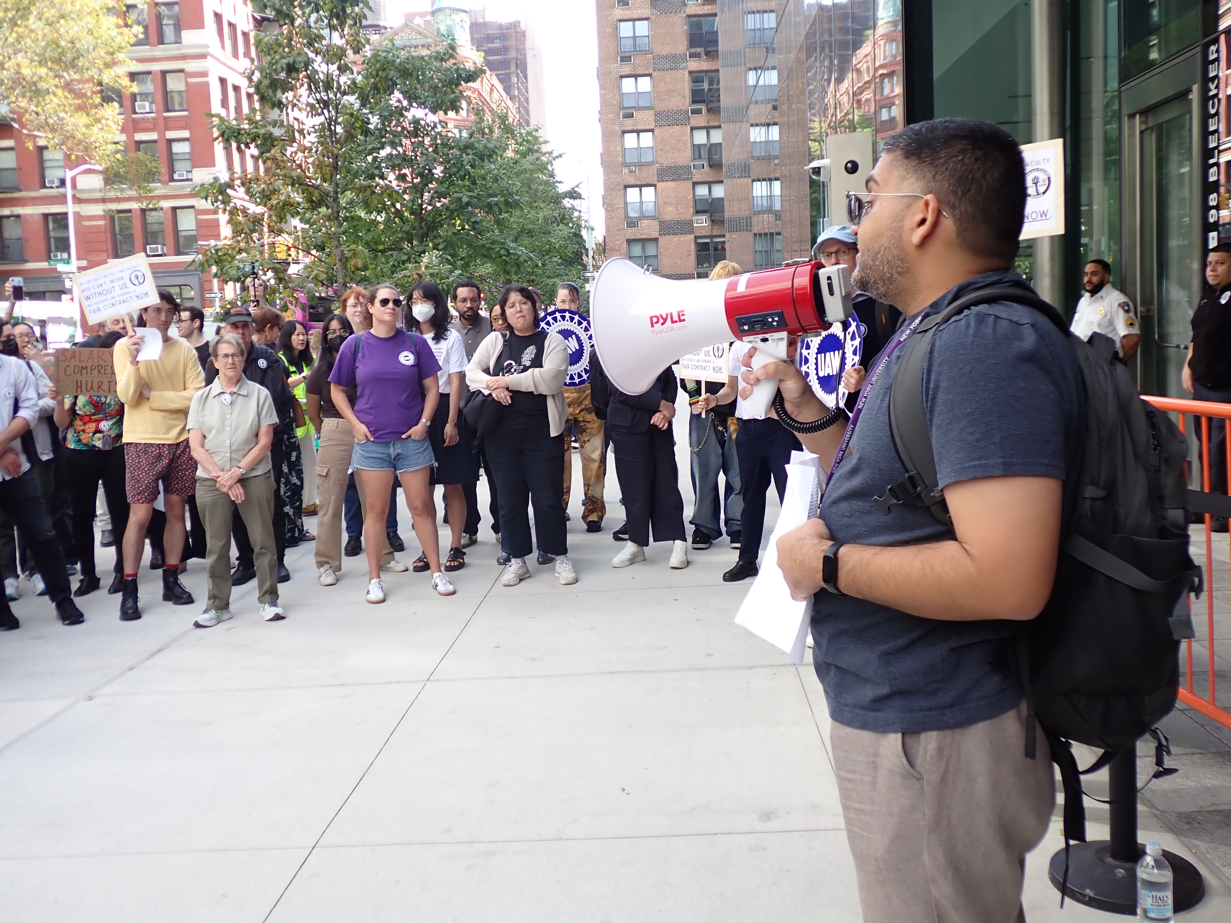 a crowd of people including NYU faculty outside, with one person holding a megaphone