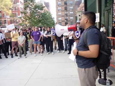 a crowd of people including NYU faculty outside, with one person holding a megaphone