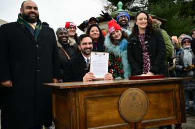 Mayor Zohran Mamdani signs an executive order Friday at Grand Army Plaza, surrounded by campaign volunteers, Tasha Van Auken, commissioner of the newly created Mayor’s Office of Mass Engagement, and Ali Najmi, chair of the Mayor’s Advisory Committee on the Judiciary